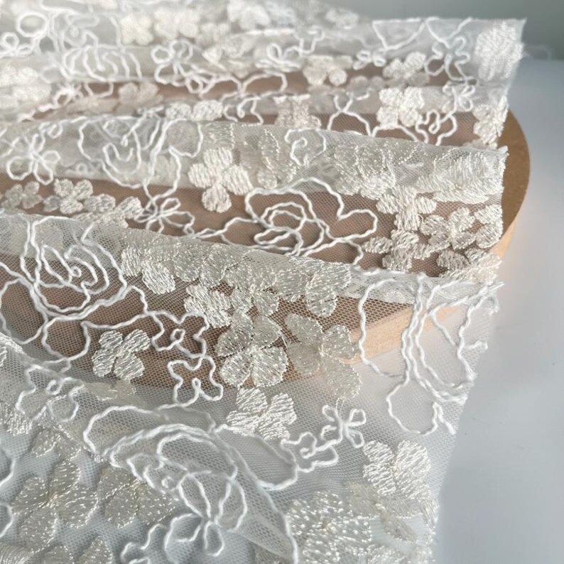 May include: White floral embroidered lace fabric with a delicate pattern. The lace is sheer and has a soft, flowing texture. The embroidery is done in a white thread and creates a delicate floral design.