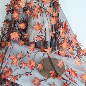 May include: Black sheer fabric decorated with embroidered maple leaves in shades of orange, red, and gold. The leaves are attached to brown branches, creating a fall-themed design. The fabric is draped over a form.