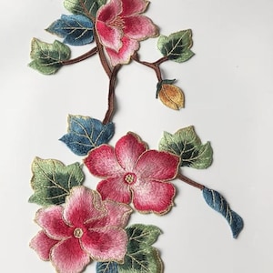Puede incluir: Aplicación floral bordada con flores rosas, hojas verdes y azules y tallos marrones. Las flores tienen un aspecto detallado y texturizado con detalles dorados. El diseño está sobre un fondo blanco, ideal para la decoración textil.