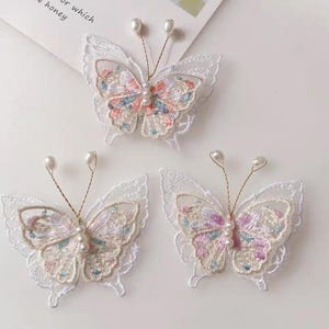 Könnte beinhalten: Drei gestickte Schmetterlingsapplikationen. Jeder Schmetterling hat zarte weiße Spitzenschwingen, bunte Blumenstickereien in Rosa, Blau und Lila und eine Perlenkette für den Körper. Die Antennen sind aus dünnem Golddraht, jede mit einer Perle an der Spitze.