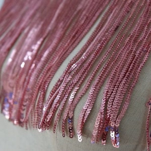 May include: Pink sequined fringe trim. The fringe is made of small, round sequins that are sewn together in a chain-like pattern. The fringe is about 1 inch long.
