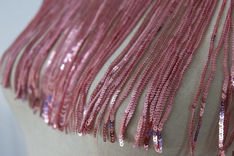 May include: Pink sequined fringe trim. The fringe is made of small, round sequins that are sewn together in a chain-like pattern. The fringe is about 1 inch long.