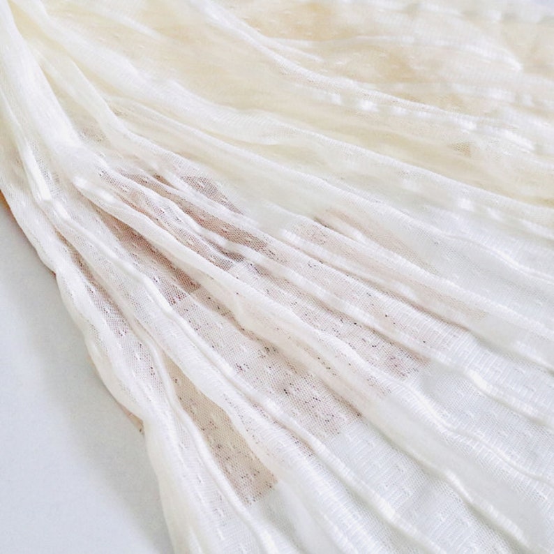 May include: White sheer fabric with a textured, dotted pattern. The fabric is pleated and has a soft, delicate look.