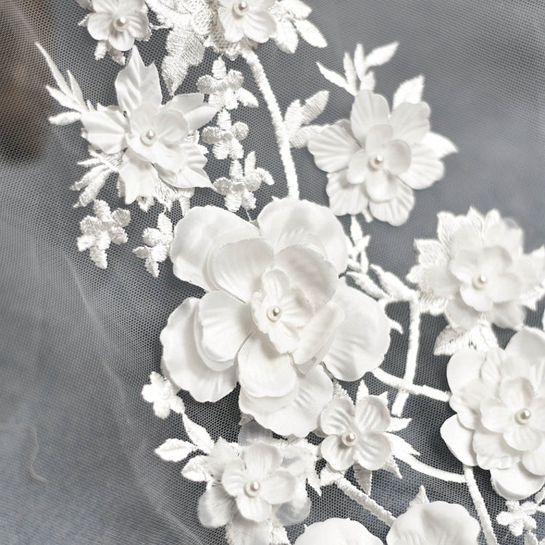 May include: White floral lace fabric with 3D flowers and pearl accents. The fabric is delicate and intricate, with a soft, feminine feel.