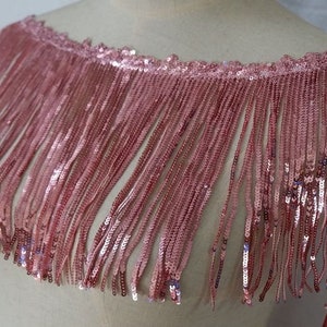 May include: Pink sequined fringe trim with a scalloped edge. The fringe is made of small sequins and is about 2 inches long.
