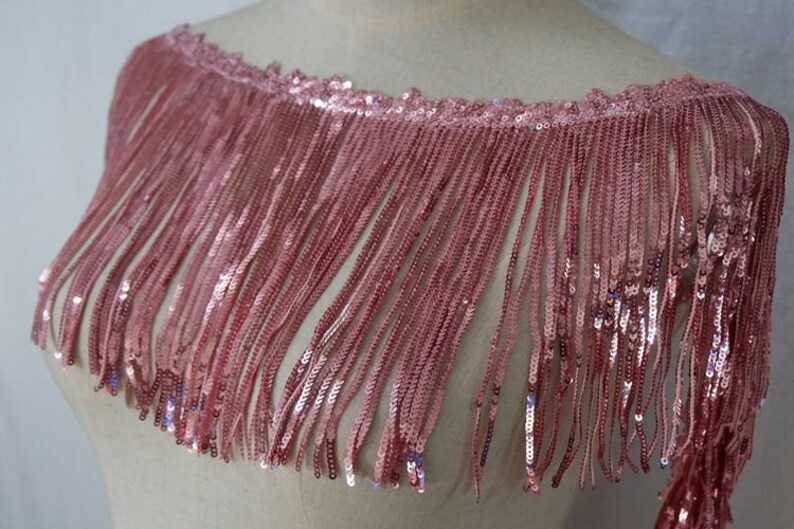 May include: Pink sequined fringe trim with a scalloped edge. The fringe is made of small sequins and is about 2 inches long.
