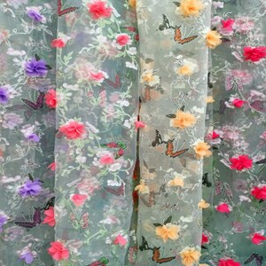 Könnte beinhalten: Ein durchsichtiger Stoff mit einem Blumenmuster und 3D-Blumenverzierungen. Der Stoff ist weiß mit rosa, lila und gelben Blumen. Die Blumen sind aus einem weichen, strukturierten Material gefertigt und mit einem zarten Stich am Stoff befestigt.