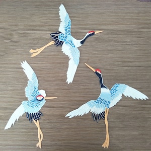 Könnte beinhalten: Drei gestickte Kranich-Applikationen in Blau und Weiß mit roten und schwarzen Akzenten. Die Kraniche sind im Flug und in stehender Pose.