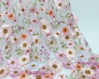 May include: Embroidered floral fabric in soft pink, peach, and lavender hues. The design features detailed flower centers and green stems and leaves, set against a sheer white backdrop. Suitable for dresses, skirts, and other clothing items. The fabric is ideal for crafting.
