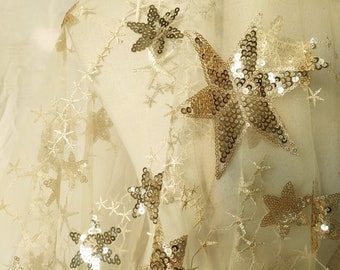 May include: Beige tulle fabric with gold star-shaped sequins. The stars are scattered across the fabric and are of varying sizes.