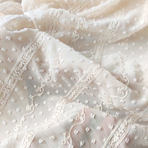 May include: White sheer fabric with a textured, dotted pattern and delicate embroidered lace trim. The fabric is soft and lightweight, perfect for creating garments or home decor.