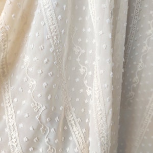 May include: White sheer fabric with a textured, dotted pattern and embroidered floral designs. The fabric has a delicate, airy feel.