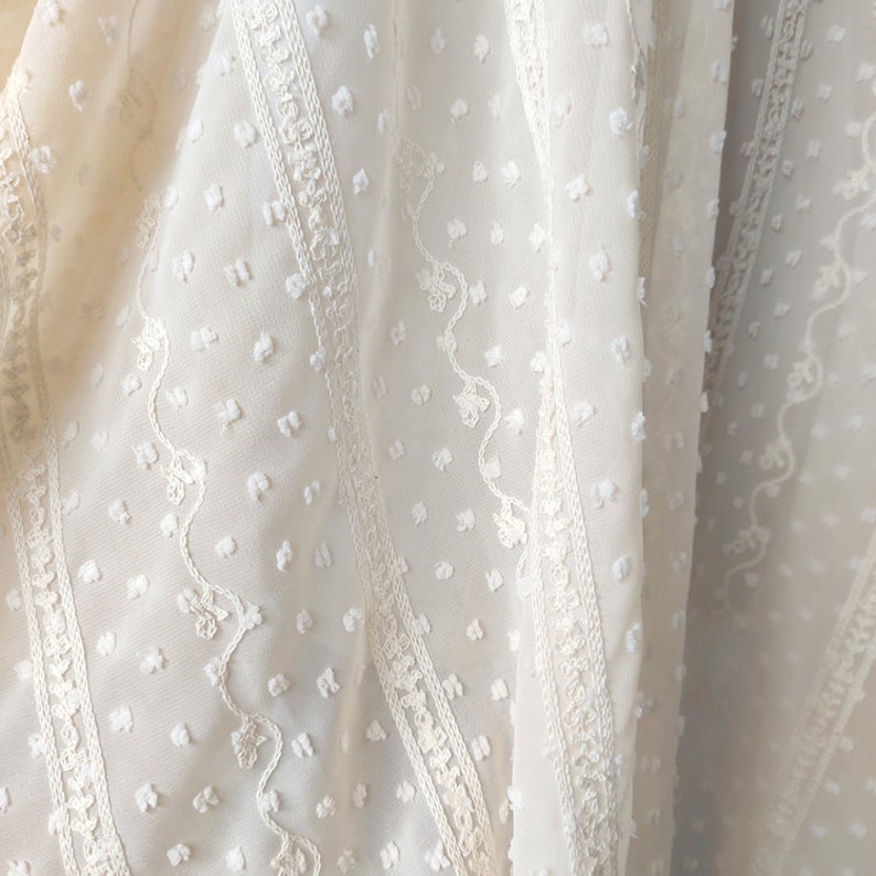 May include: White sheer fabric with a textured, dotted pattern and embroidered floral designs. The fabric has a delicate, airy feel.