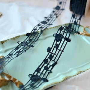 May include: Black ribbon with a musical note design. The ribbon is on a light blue surface.
