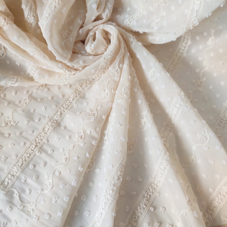 May include: A close-up of a cream-colored sheer fabric with a delicate embroidered pattern of small dots and floral designs. The fabric has a textured, slightly raised surface.