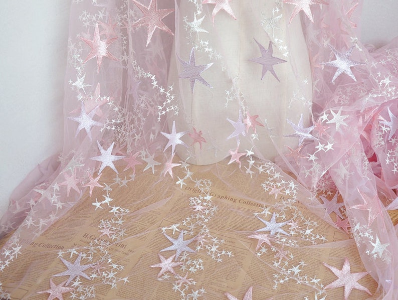 May include: Pink tulle fabric with white and pink embroidered stars. The fabric is sheer and delicate, and the stars are scattered randomly across the surface.