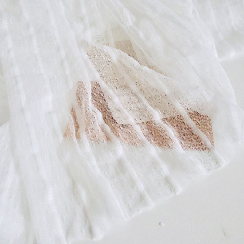 May include: White sheer fabric with a textured, dotted pattern. The fabric is pleated and has a soft, delicate feel.