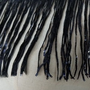 May include: Black fringe trim with sequins. The fringe is made of thin, black threads and is about 1 inch long. The sequins are small and scattered throughout the fringe.