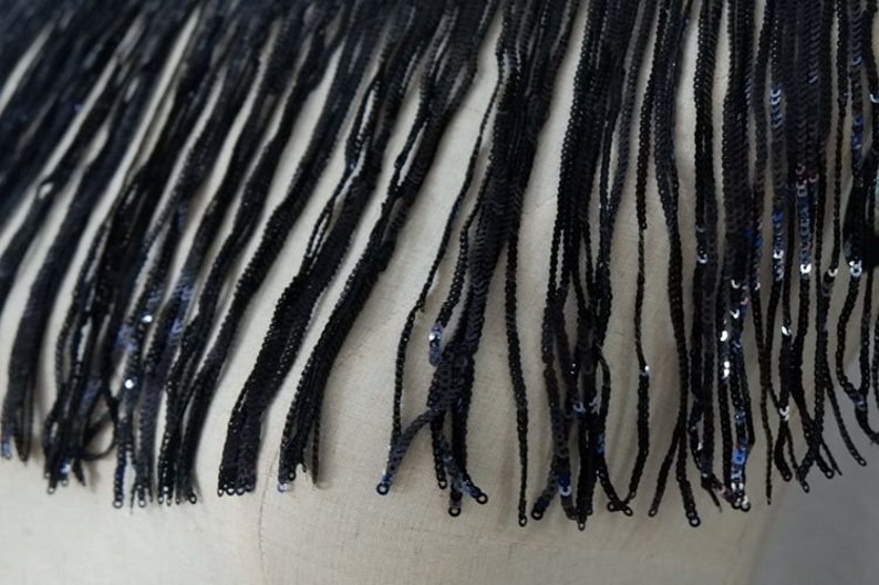 May include: Black fringe trim with sequins. The fringe is made of thin, black threads and is about 1 inch long. The sequins are small and scattered throughout the fringe.