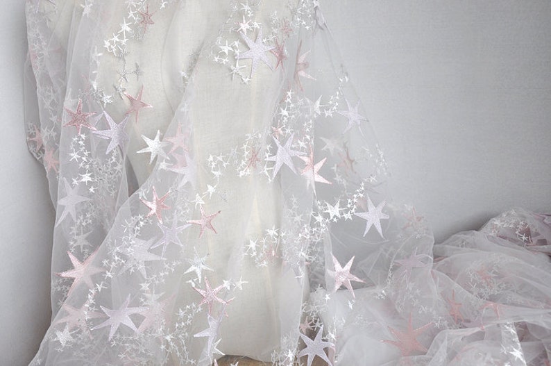 May include: White tulle fabric with pink embroidered stars. The fabric is sheer and delicate, perfect for crafting or adding a touch of sparkle to any project.