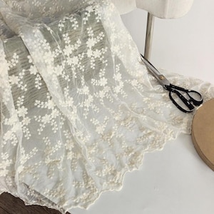 Könnte beinhalten: Ein transparenter, cremeweißer Stoff mit einem komplizierten gestickten Blumenmuster aus kleinen Blüten und rankenden Zweigen. Das Material hat einen gewellten Rand und liegt neben einer schwarzen Metallschere auf einer flachen Oberfläche.