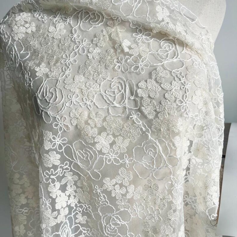 May include: White floral embroidered lace fabric with a delicate pattern of small flowers and larger roses. The lace is sheer and has a soft, flowing texture.