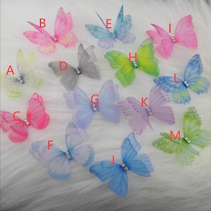 Könnte beinhalten: Eine Sammlung farbenfroher, durchsichtiger Schmetterlingsverzierungen. Die Schmetterlinge sind in verschiedenen Farbtönen erhältlich, darunter Rosa, Blau, Grün und Lila. Jeder Schmetterling hat einen kleinen, funkelnden Edelstein in der Mitte. Diese dekorativen Schmetterlinge sind ideal für Bastelprojekte.