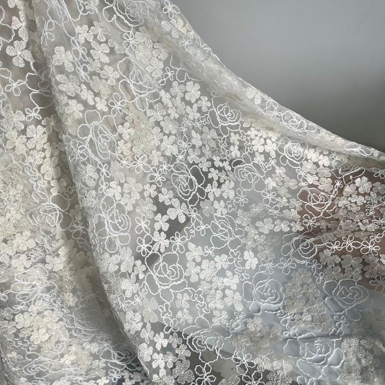 May include: White floral lace fabric with a delicate pattern of small flowers and larger roses. The lace is sheer and has a soft, flowing texture.