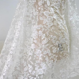 May include: White floral lace fabric with intricate embroidered flower and leaf patterns. The delicate design is visible throughout the sheer material, creating a textured and elegant look. Ideal for bridal wear or decorative projects.