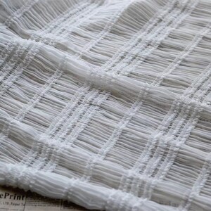May include: White, delicate, sheer fabric with a crinkled texture. The fabric is woven in a grid pattern, creating a subtle, raised design.