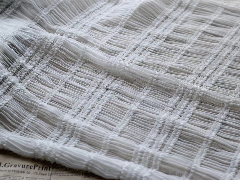 May include: White, delicate, sheer fabric with a crinkled texture. The fabric is woven in a grid pattern, creating a subtle, raised design.