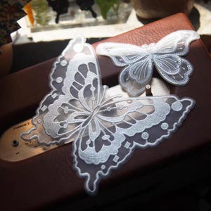 Könnte beinhalten: Drei weiße, gestickte Schmetterlingsapplikationen. Der größte Schmetterling ist teilweise sichtbar, mit komplizierten, spitzenartigen Details. Ein kleinerer Schmetterling ist darüber positioniert. Die Applikationen befinden sich auf einer braunen Oberfläche.