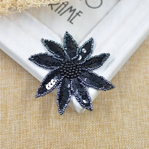 Könnte beinhalten: Schwarze Pailletten-Blumenapplikation mit einem Perlenkern. Die Blume hat 10 Blütenblätter und ist etwa 5 cm im Durchmesser.