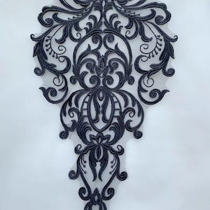 Könnte beinhalten: Eine schwarze, gestickte Spitzenapplikation mit einem verzierten, symmetrischen floralen Design. Die Applikation weist wirbelnde Muster und filigrane Details auf, ideal zum Verzieren von Kleidungsstücken oder Bastelarbeiten. Die Spitze ist vor einem weißen Hintergrund platziert.