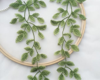 May include: Two green embroidered leafy vines with a light green color gradient. The vines are displayed on a wooden embroidery hoop.