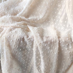May include: Close-up of a cream-colored sheer fabric with a textured dot pattern and delicate embroidered stripes.