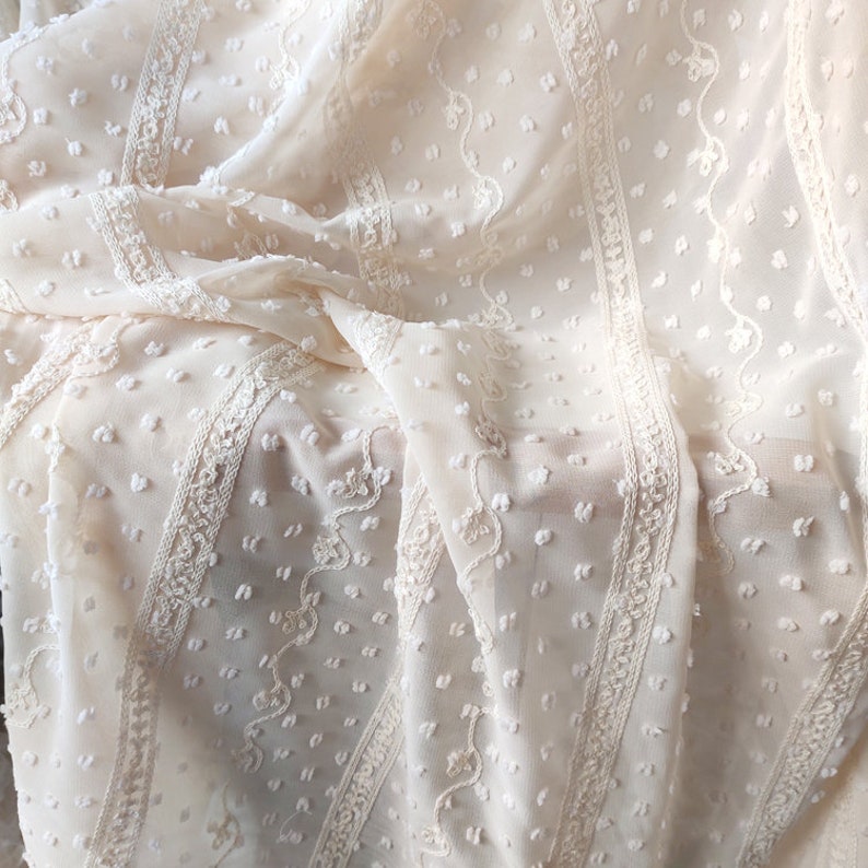 May include: Close-up of a cream-colored sheer fabric with a textured dot pattern and delicate embroidered stripes.