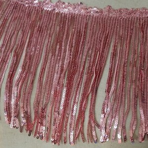 May include: Pink sequined fringe trim with a scalloped edge. The fringe is made of small, shiny sequins that are sewn together in a continuous line.