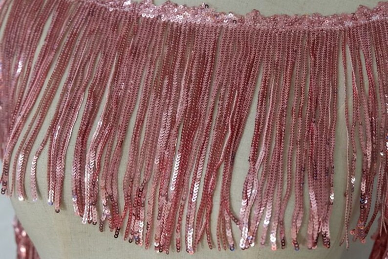 May include: Pink sequined fringe trim with a scalloped edge. The fringe is made of small, shiny sequins that are sewn together in a continuous line.