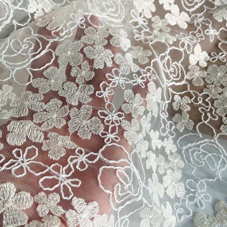 May include: White sheer fabric with silver floral embroidery. The fabric is delicate and has a soft, romantic feel.