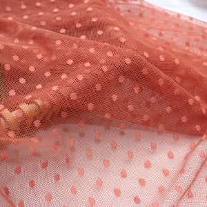 May include: A close-up of a rust-colored tulle fabric with small white polka dots. The fabric is soft and delicate, and it has a slightly shimmery finish.