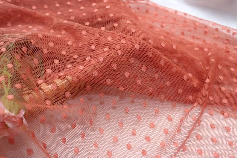 May include: A close-up of a rust-colored tulle fabric with small white polka dots. The fabric is soft and delicate, and it has a slightly shimmery finish.