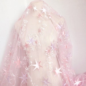 May include: Pink tulle fabric with embroidered white and pink stars. The fabric is sheer and delicate, perfect for crafting or adding a touch of whimsy to a project.