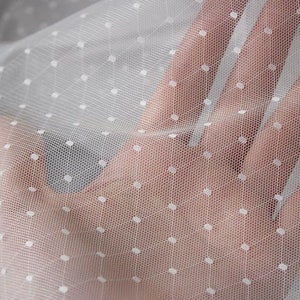 May include: Close-up of white tulle fabric with small, evenly spaced white polka dots. The sheer material is held up, showing the texture and transparency of the fabric. Ideal for crafting, bridal wear, or decorative projects.