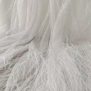 May include: White tulle fabric with a delicate fringe and a subtle sparkle. The fabric is sheer and lightweight, making it ideal for creating a variety of garments and accessories.