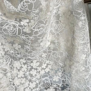 May include: White floral lace fabric with a delicate pattern of small flowers and larger roses. The lace is sheer and has a soft, romantic feel.