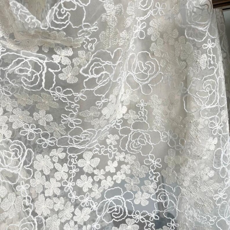May include: White floral lace fabric with a delicate pattern of small flowers and larger roses. The lace is sheer and has a soft, romantic feel.