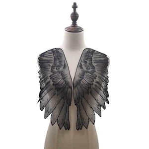 Puede incluir: Alas de ángel de encaje negro exhibidas en un maniquí. Las alas presentan patrones de plumas detallados y están diseñadas para ser usadas sobre los hombros. El encaje delicado crea un efecto semitransparente.