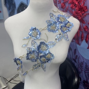 Könnte beinhalten: Blumenapplikation mit blauen Paillettenblumen, goldenen Zentren und goldenen Fäden. Das Design befindet sich auf einem transparenten, weißen Mesh-Träger. Ideal zum Verzieren von Kleidung oder Accessoires, für einen Hauch von Eleganz.