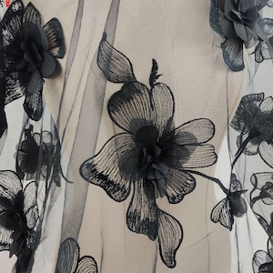 Puede incluir: Tela de encaje floral negro con adornos de flores en 3D. La tela presenta grandes flores bordadas detalladas en negro, con delicados pétalos y un fondo transparente. Ideal para vestidos de novia, vestidos de noche o proyectos decorativos.